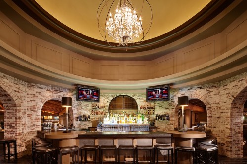Elegant bar with a chandelier, brick walls, and two screens displaying drinks, surrounded by high stools.