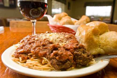 A plate of spaghetti topped with meatballs and marinara sauce, served with rolls and a glass of red wine.