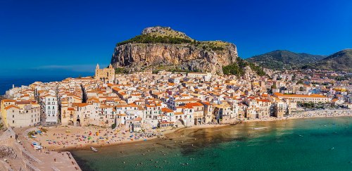 Aerial view of a coastal town with white buildings and orange rooftops, set against a rocky hill and clear blue water.