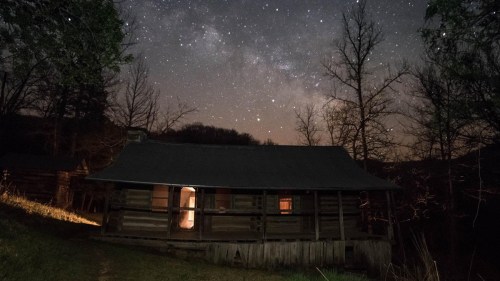 A rustic cabin illuminated by warm light under a starry night sky, surrounded by trees and a serene landscape.