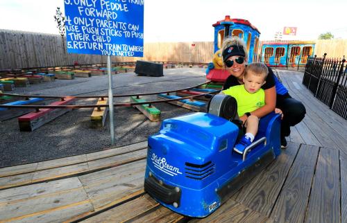 A woman and a young boy sit on a blue toy train at a play area, with a sign listing rules in the background.