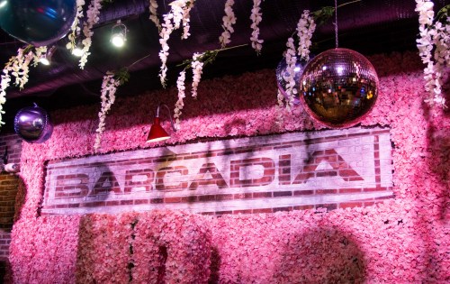 A vibrant wall covered in pink flowers with a sign reading "BARCADIA," adorned with disco balls and hanging decorations.
