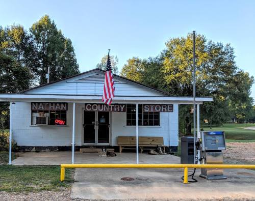 A small country store with a flag, sign reading "Nathan Country Store," and a gas pump outside. Trees in the background.