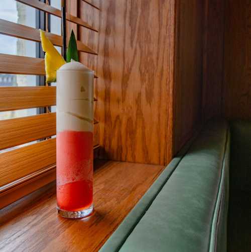 A tall cocktail glass with layered red and white drinks, garnished with a pineapple slice and green leaves, on a wooden table.