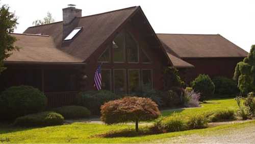 A large, rustic house with a triangular window, surrounded by greenery and a small American flag in the front yard.