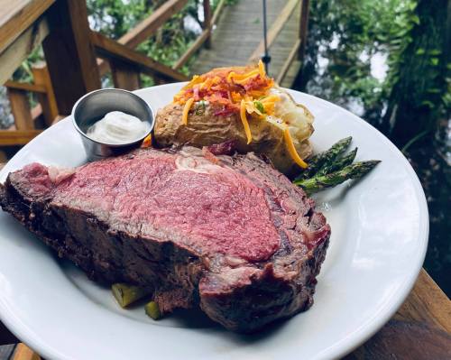 A plate featuring a juicy prime rib, baked potato with toppings, and asparagus, set against a lush outdoor backdrop.