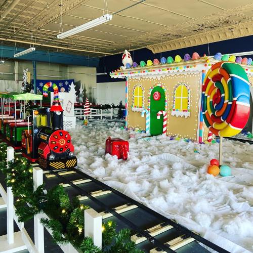 A colorful gingerbread house with a train, surrounded by fake snow and festive decorations in a large indoor space.