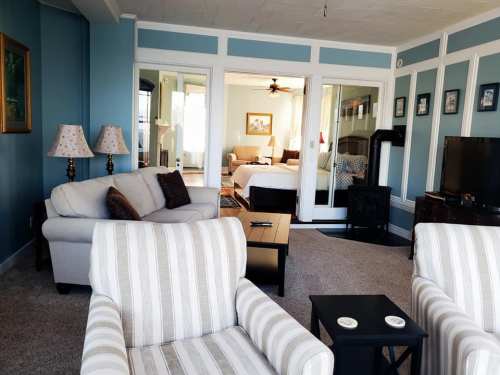 A cozy living room with blue walls, a sofa, striped chairs, and a view of a bedroom through glass doors.