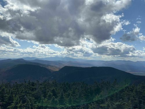 A panoramic view of mountains under a cloudy sky, with lush green trees in the foreground.