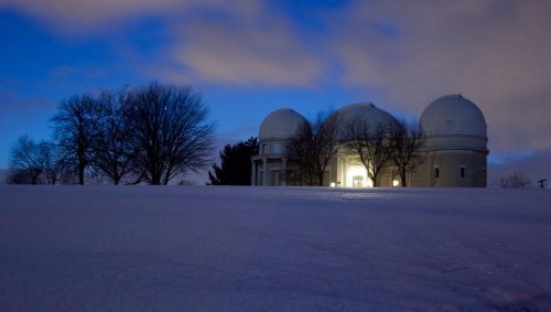 A snowy landscape at dusk featuring a large observatory with three domes and bare trees in the foreground.