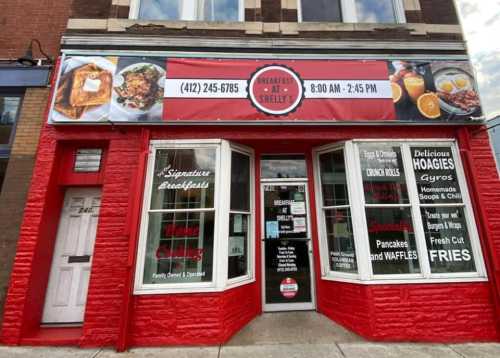 Exterior of a restaurant with a red facade, featuring a sign with the name, hours, and menu items like breakfast and hoagies.