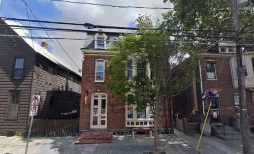 A brick house with a peaked roof, surrounded by trees and power lines, next to similar buildings on a street.