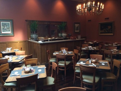 Cozy restaurant interior with wooden tables, chairs, and warm lighting, featuring a bar area and decorative plants.