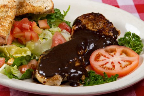 A plate featuring grilled chicken with dark sauce, fresh salad, sliced tomatoes, and toasted bread on a checkered tablecloth.
