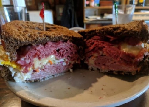 A close-up of a hearty sandwich with layers of meat, cheese, and egg, served on dark rye bread with toothpicks.