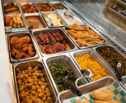 A variety of Southern-style dishes in metal trays, including fried chicken, greens, corn, and sausage.