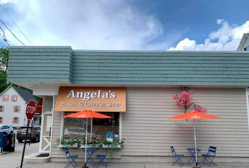 Exterior of Angela's Pasta & Cheese Shop, featuring orange umbrellas and a stop sign nearby under a blue sky.