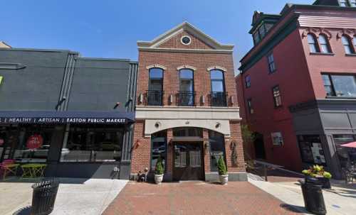 A brick building with a decorative facade, flanked by two colorful storefronts on a sunny day.