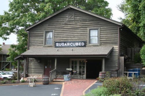 A rustic wooden building with a sign reading "SUGAR CUBED" and a welcoming entrance, surrounded by greenery.