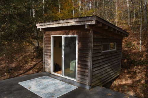 A small wooden cabin with a glass door, surrounded by trees and autumn foliage. A rug is placed outside on the concrete.