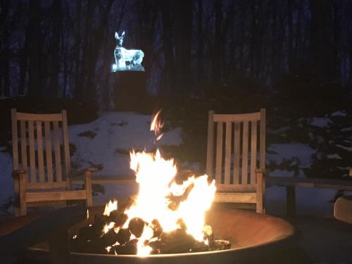 A cozy fire pit with flames, two wooden chairs, and a glowing deer statue in a snowy, dark forest setting.