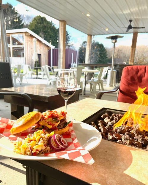 A burger with toppings and a side of salad, paired with a glass of red wine, next to a fire pit on a patio.