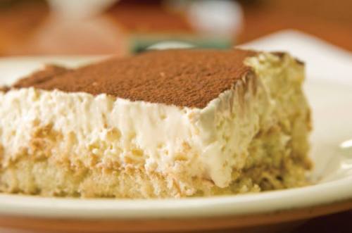 A slice of tiramisu on a white plate, featuring layers of coffee-soaked cake and creamy mascarpone topped with cocoa.