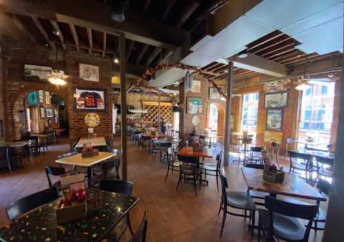 Interior of a rustic restaurant with wooden beams, brick walls, and tables set for dining. Bright windows let in natural light.