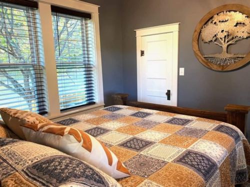 Cozy bedroom with a quilted bedspread, wooden accents, and a decorative tree wall art. Natural light from windows.