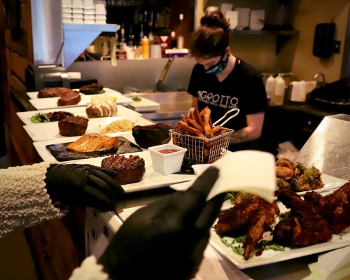 A variety of dishes including meats and sides are prepared in a restaurant kitchen, with a masked worker in the background.