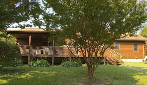 A wooden house with a front porch surrounded by greenery and trees on a sunny day.
