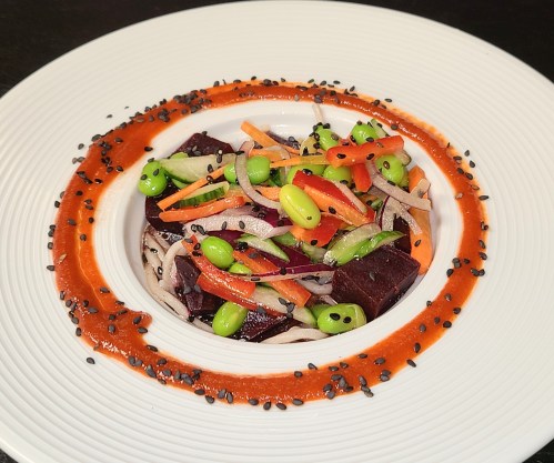 A colorful dish featuring noodles, beets, edamame, and vegetables, garnished with a red sauce and black sesame seeds.