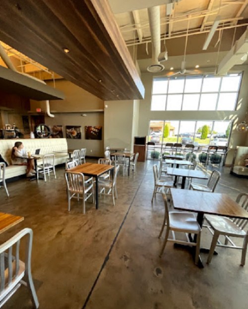 A spacious café interior with wooden tables, metal chairs, and large windows, featuring a person working on a laptop.