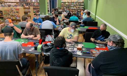 A group of people playing card games in a game store, wearing masks and seated at tables with game materials.