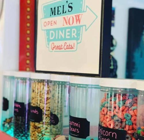 A colorful diner sign reads "Mel's Open Now" with jars of candy, including gummy bears and colorful cereal.