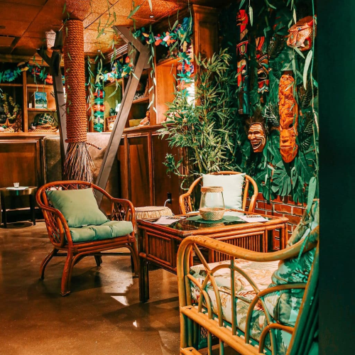 A cozy, tropical-themed room with wooden furniture, greenery, and decorative masks on the walls.