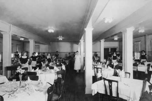 A vintage dining room with neatly set tables and waitstaff in uniforms, showcasing a classic restaurant atmosphere.