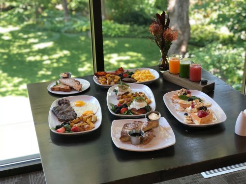 A table with various breakfast dishes, including eggs, steak, pancakes, and fresh fruit, set against a green garden view.