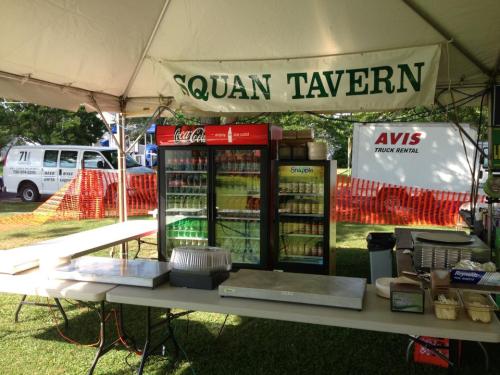 A food stand under a tent with a sign reading "Squan Tavern," featuring drinks and supplies on tables.