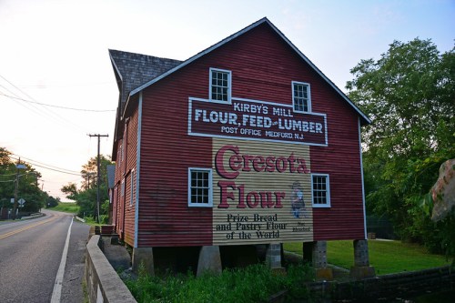 Red barn-style building with signs for flour, feed, and lumber, located by a road in Medford, NJ.