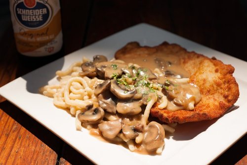 A plate of breaded chicken with creamy mushroom sauce over pasta, garnished with herbs, next to a beer bottle.