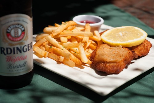 A plate of golden fries, two fried fish fillets with a lemon slice, and a small bowl of ketchup, alongside a beer.