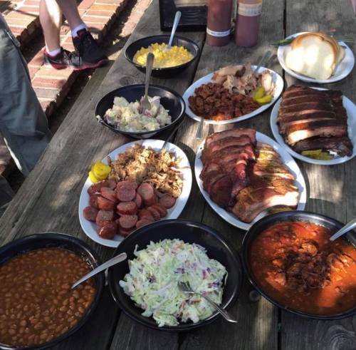 A spread of barbecue dishes on a wooden table, including meats, sides, and sauces.