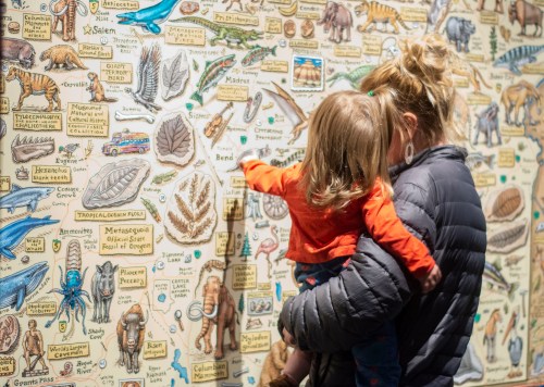A woman holds a child as they explore a colorful wall covered in illustrations of animals and plants.
