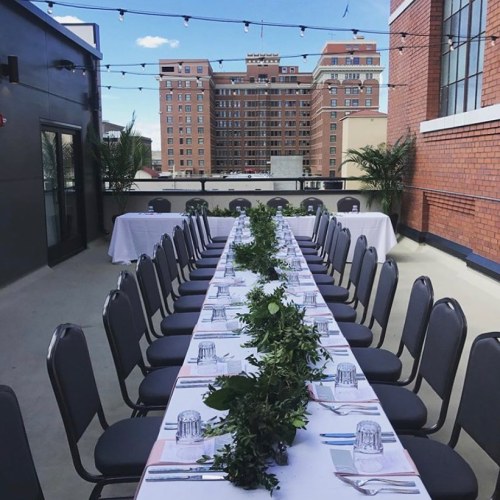 A long dining table set with plates and glasses, decorated with greenery, on a rooftop with city buildings in the background.