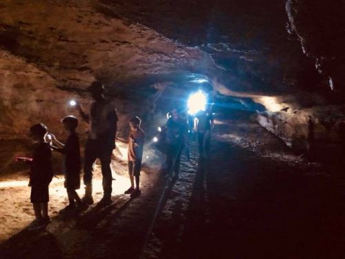 A group of children and adults explore a dark cave, illuminated by flashlights and a bright light in the background.