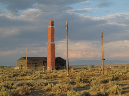 An old building with a tall red chimney stands in a desert landscape under a cloudy sky. Sagebrush surrounds the area.