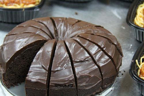 A round chocolate cake with a glossy frosting, sliced into even pieces, placed on a table with other food items.