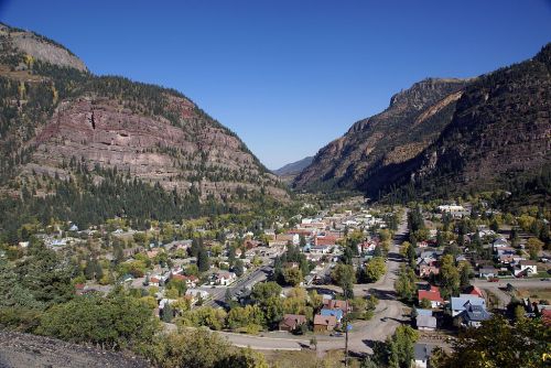 A scenic view of a small town nestled between mountains, surrounded by trees and clear blue skies.