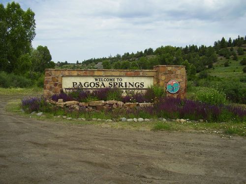 Welcome sign for Pagosa Springs, surrounded by greenery and flowers, with a cloudy sky in the background.